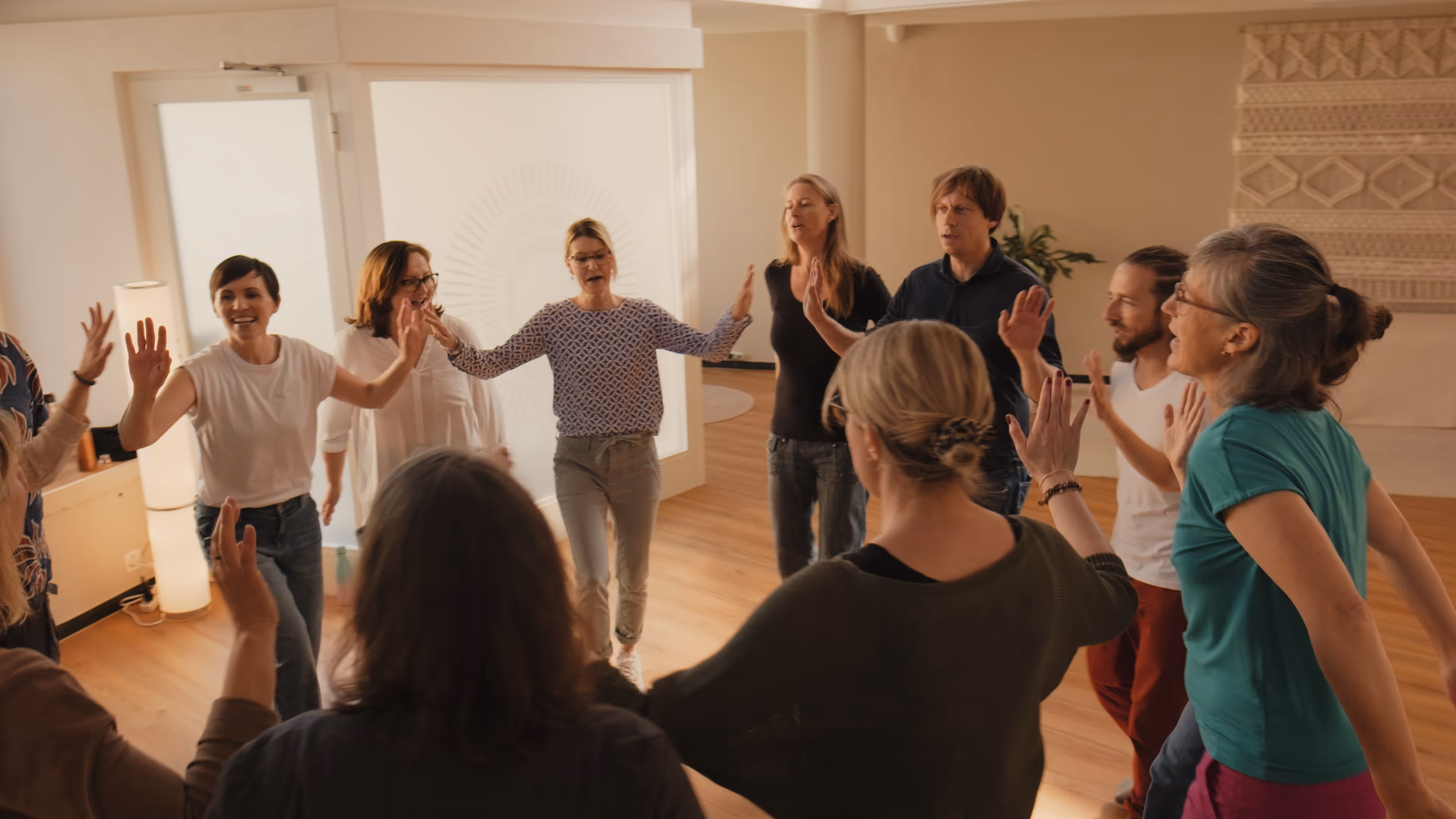 Eine Gruppe von Menschen steht im Kreis in einem Raum und hebt die Hände, einige lächeln und wirken fröhlich.