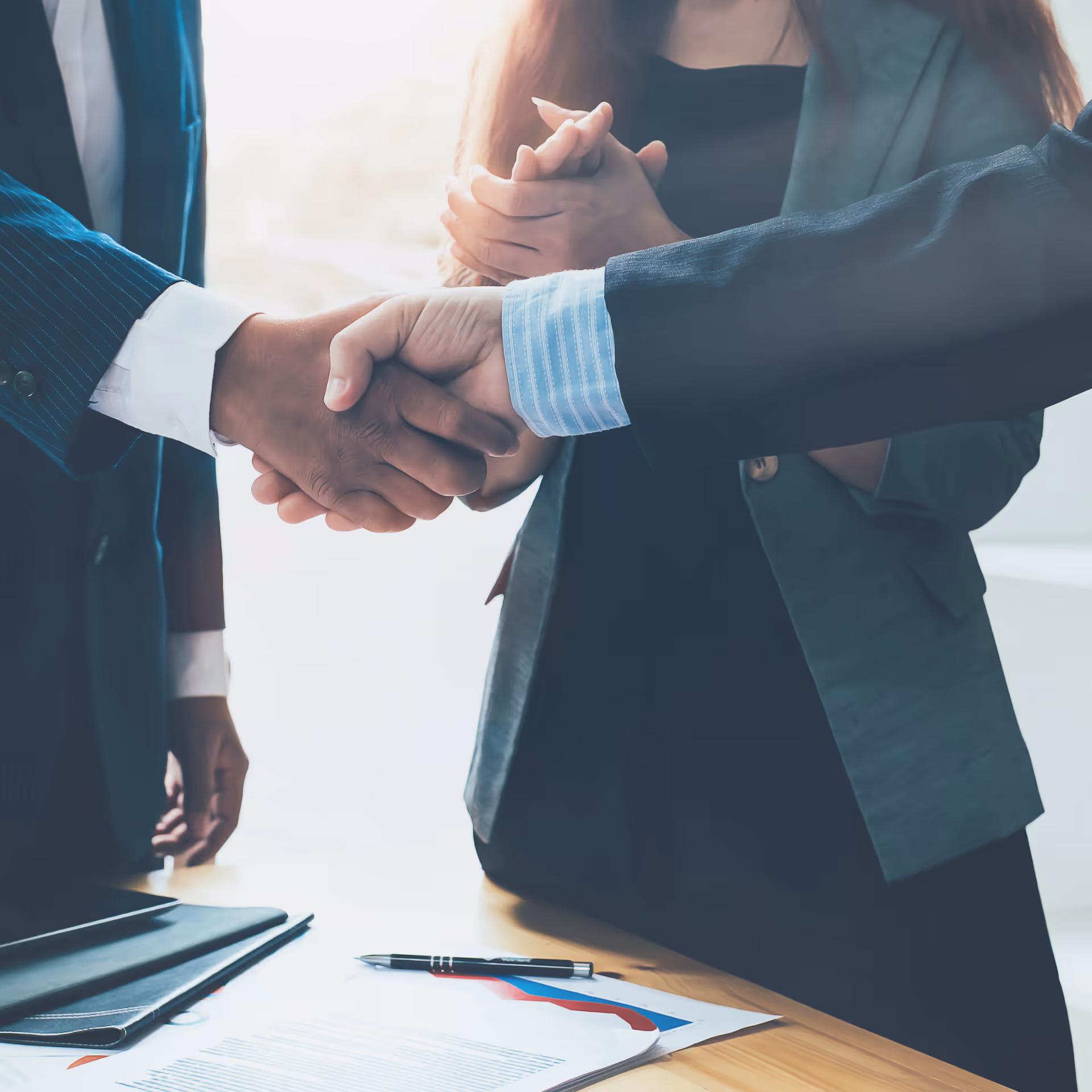 Two businesspeople shaking hands over a desk with charts and a pen, while a woman claps in the background.