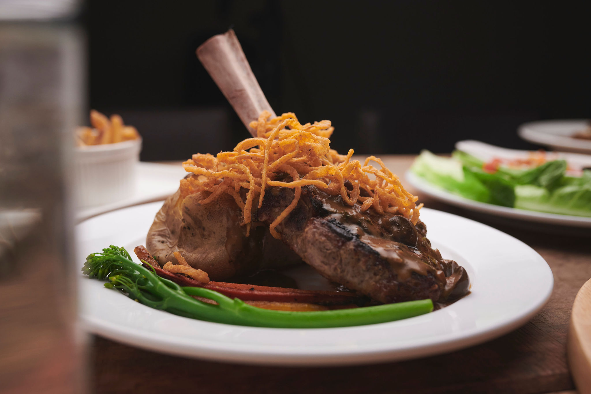 A bone-in lamb chop upright on an angle in the middle of a white circular plate, with vegetables on the side.