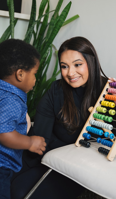 Woman smiling and engaging with a young boy while holding a colorful wooden abacus in a room with green plants.