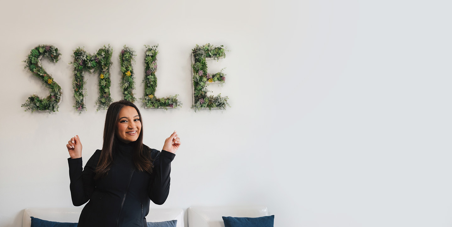 Smiling woman in black standing in front of a white wall with the word 'SMILE' spelled out in greenery above her.