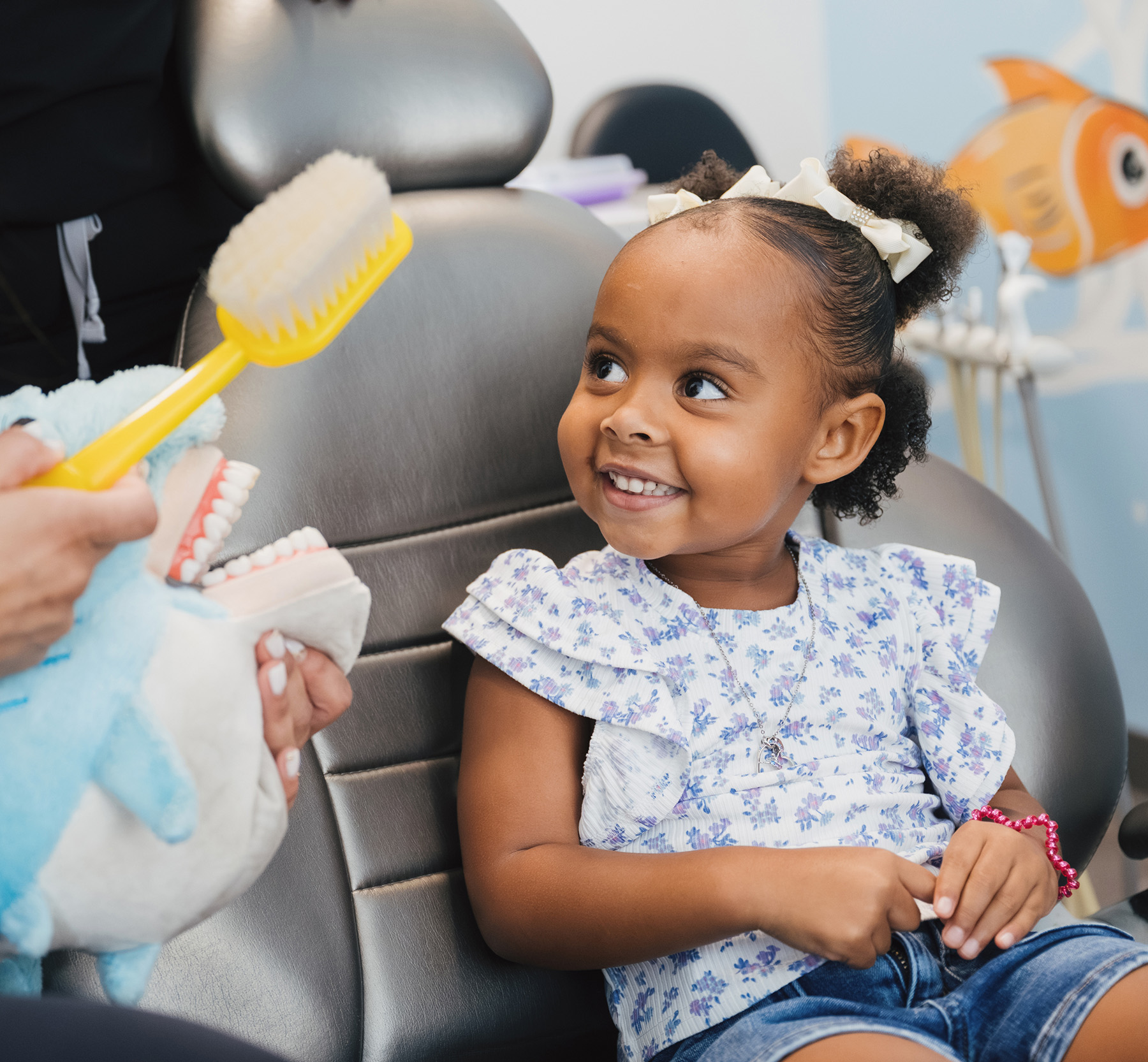 Smiling toddler girl sits in a dental chair looking at a person holding a blue shark puppet and a large yellow toothbrush.