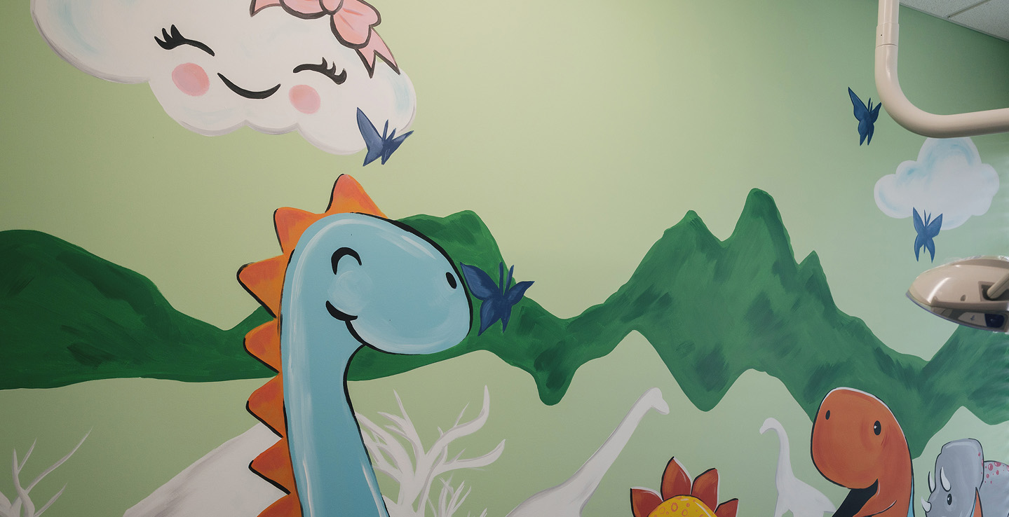 Colorful mural of smiling cartoon dinosaurs, green mountains, butterflies, and a smiling cloud with a pink bow on a light green wall.