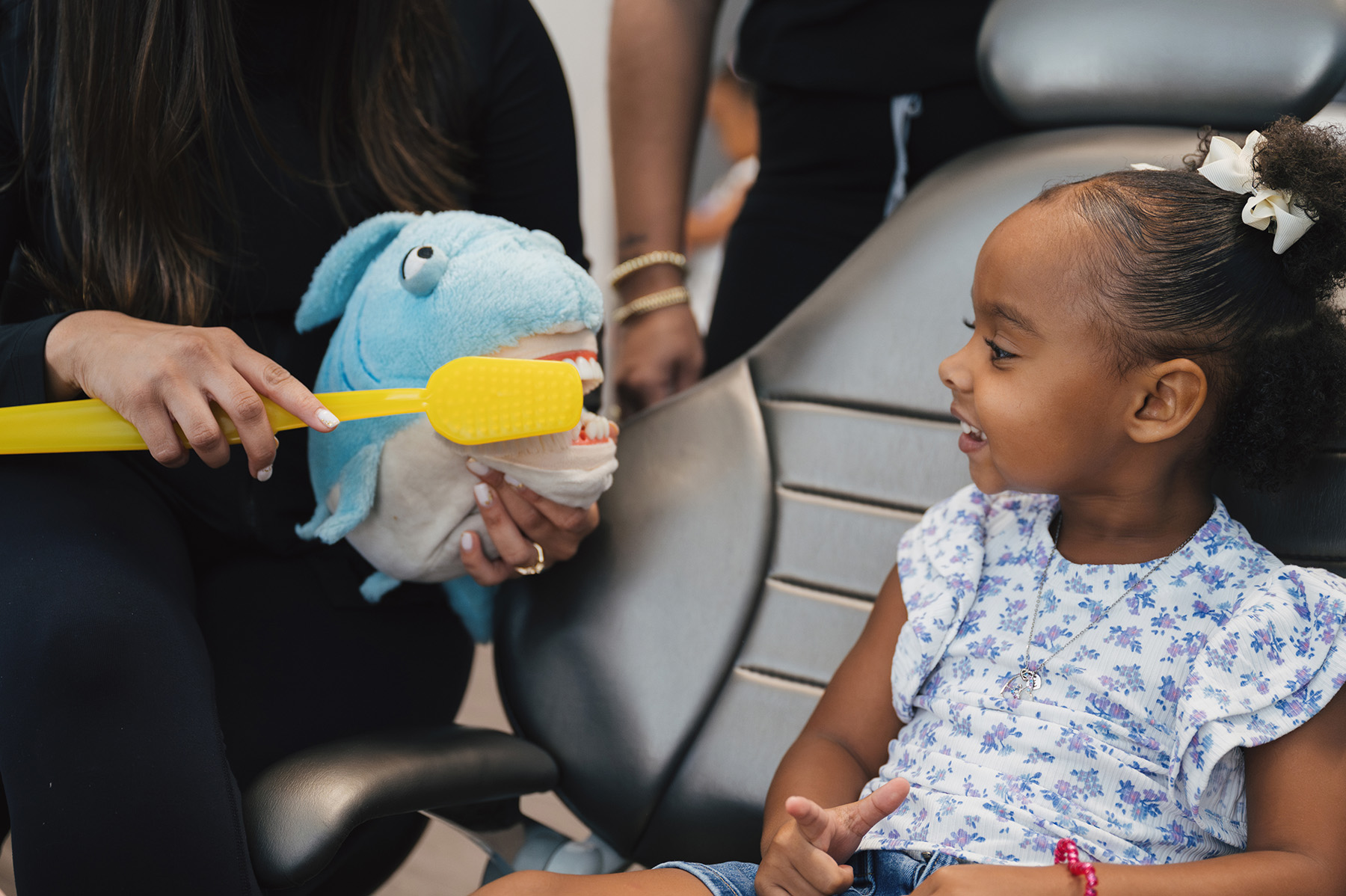 Smiling young girl watches an adult demonstrating tooth brushing on a blue shark puppet with a large yellow toothbrush.