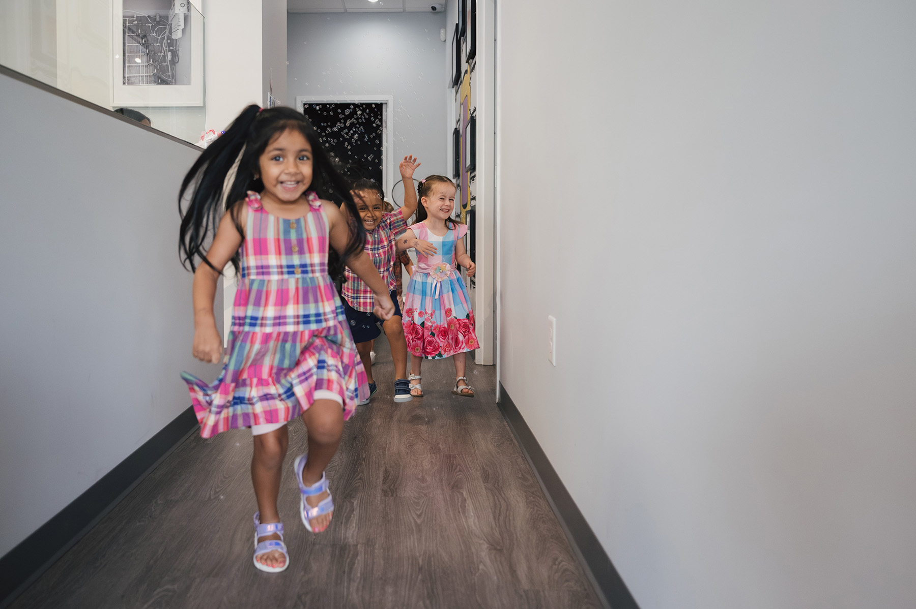 Three smiling children running down a hallway, with the child in front wearing a pink plaid dress.