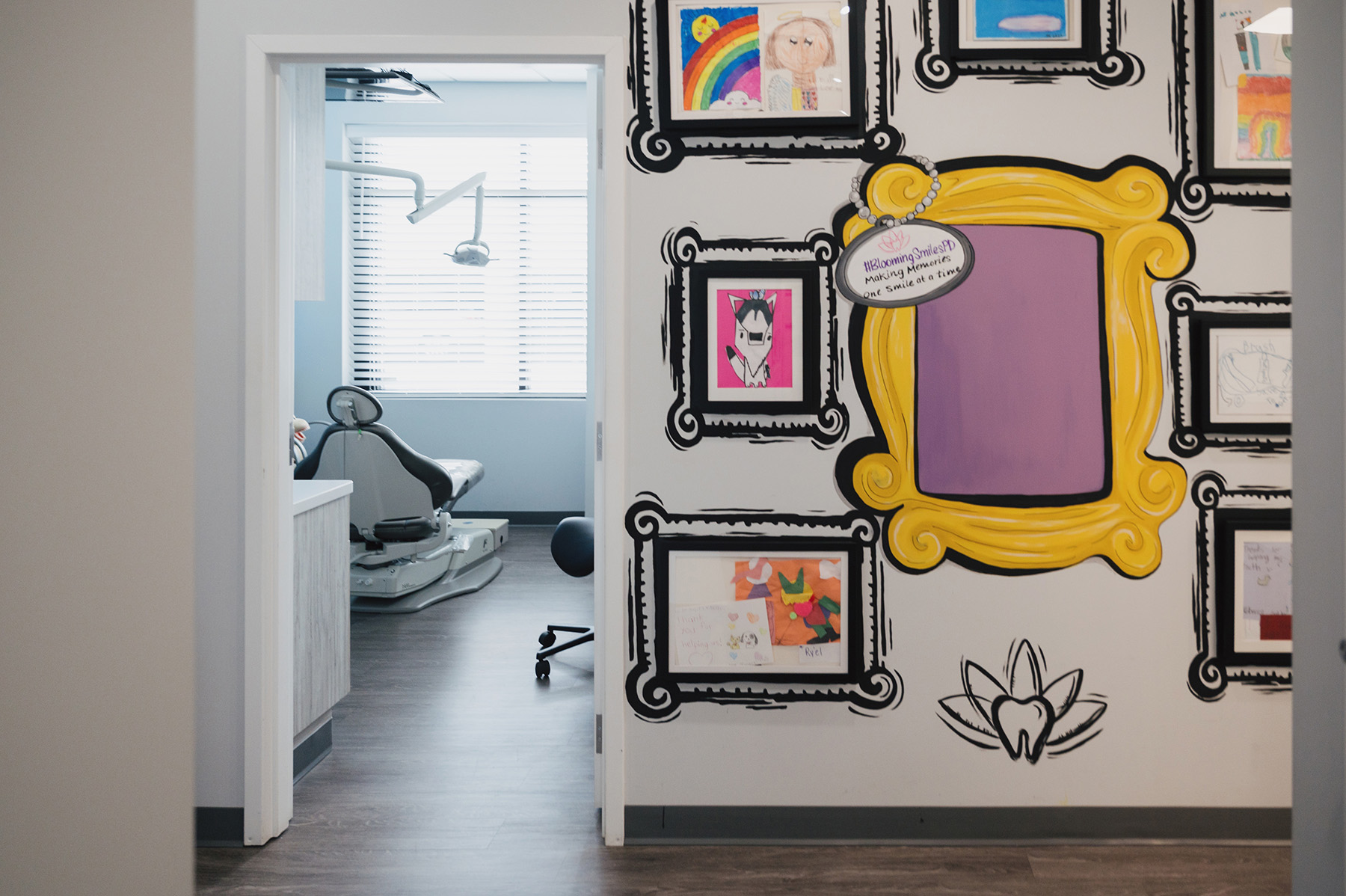Dental examination room with a black dental chair, overhead lamp, and a wall decorated with colorful children's drawings in black frames.