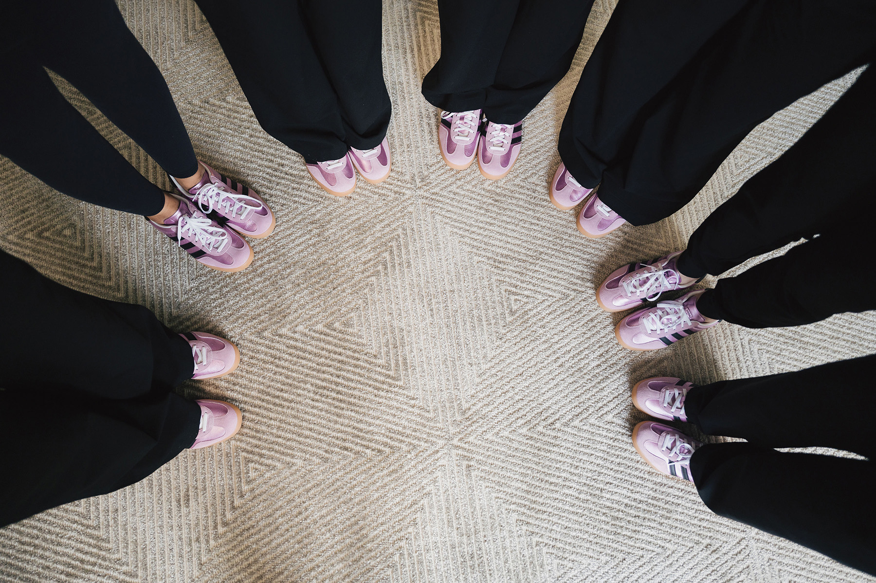 Top-down view of six people wearing similar pink and purple sneakers standing in a circle on a patterned carpet.