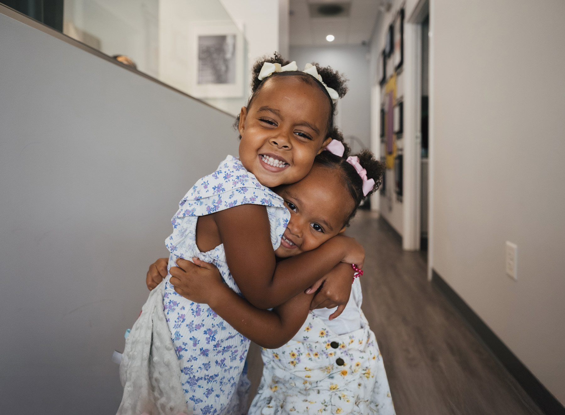 Two young girls with floral dresses and bows hugging and smiling in a hallway.
