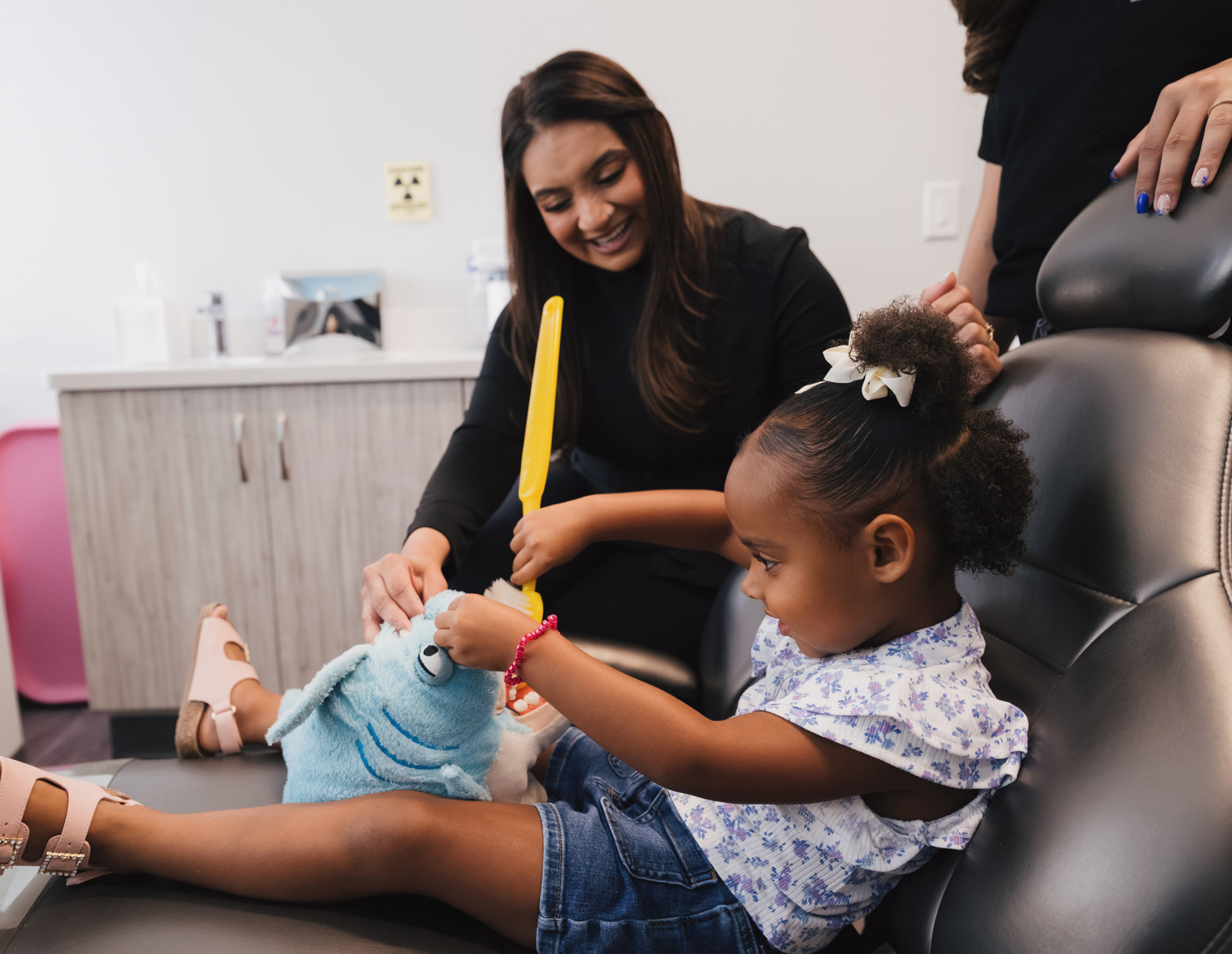 Young girl sitting in a dental chair playing with a shark puppet while a smiling dental professional holds a large toothbrush.