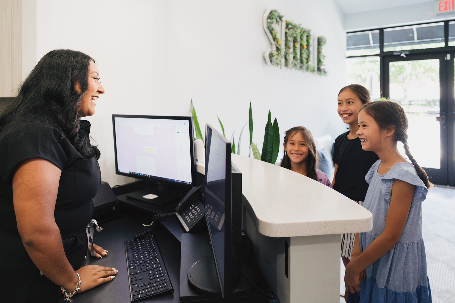 Smiling receptionist talking to three smiling young girls standing at a reception desk in a bright, modern office.