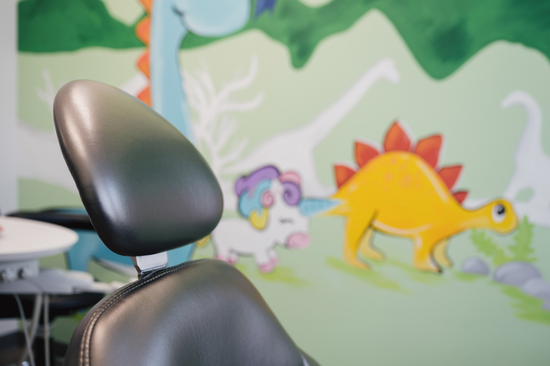 Close-up of a black dental chair with a colorful mural of cartoon dinosaurs on the wall in the background.