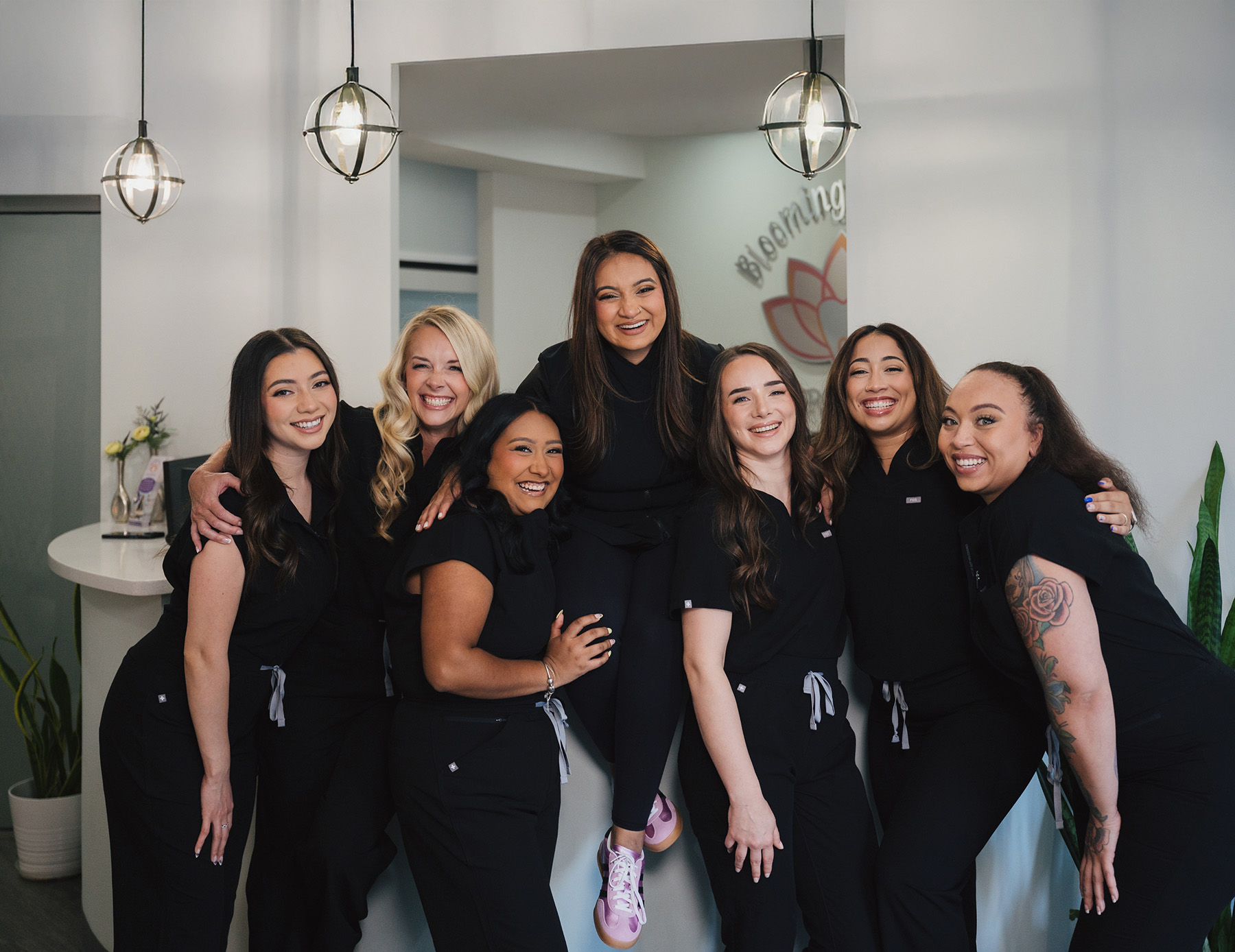 Group of seven smiling women in black scrubs posing together indoors under hanging lights.