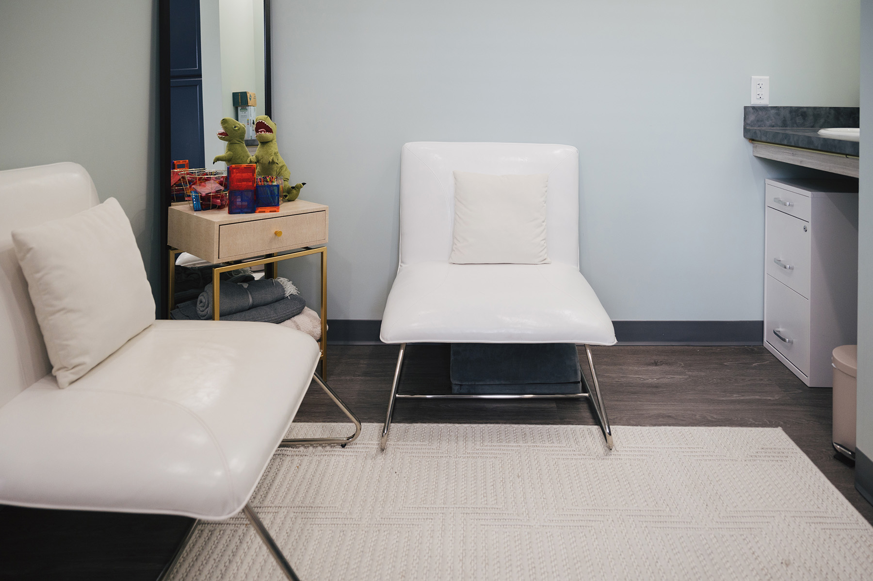 Minimalist seating area with two white chairs, each with a pillow, a small side table holding dinosaur toys and blocks, and a textured white rug on a dark floor.