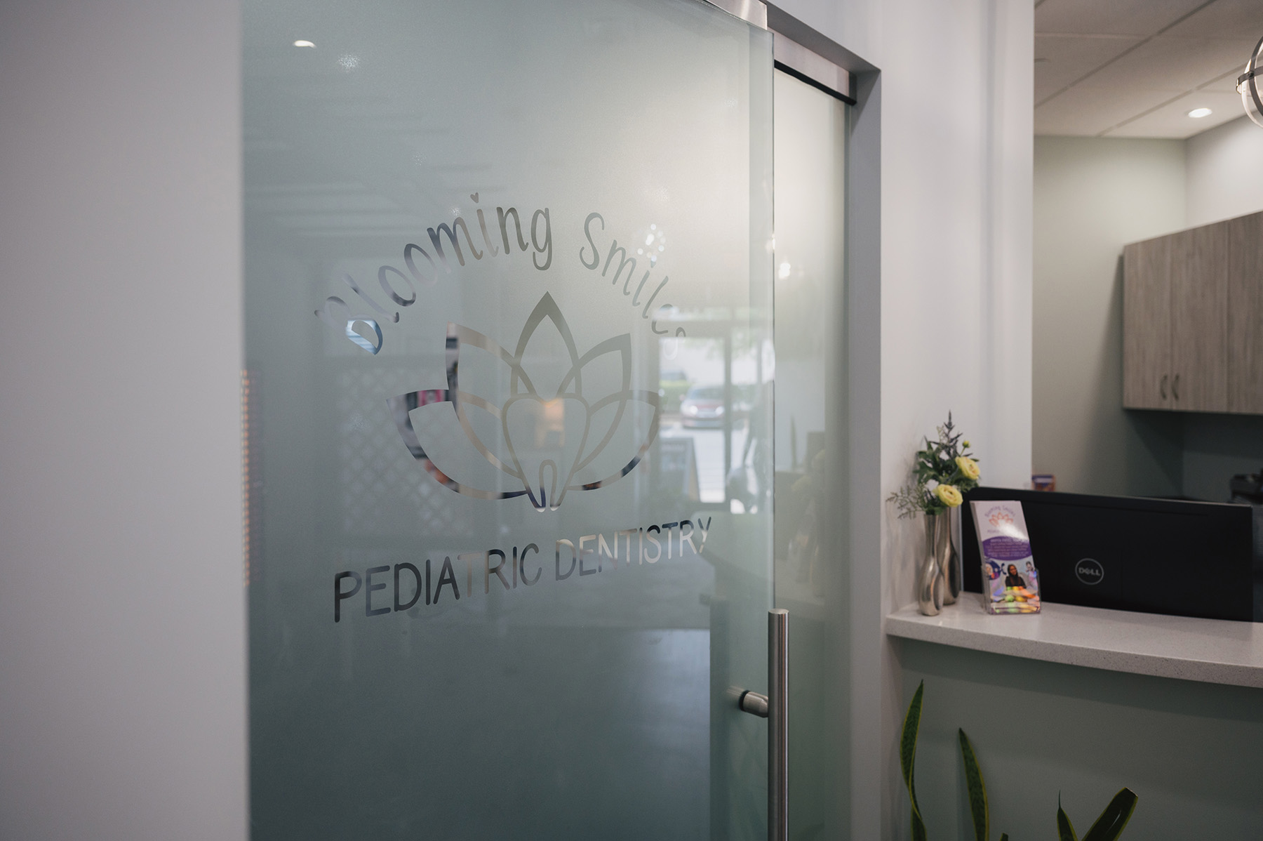Frosted glass door with 'Blooming Smiles Pediatric Dentistry' logo next to a reception desk with flowers and a computer monitor.