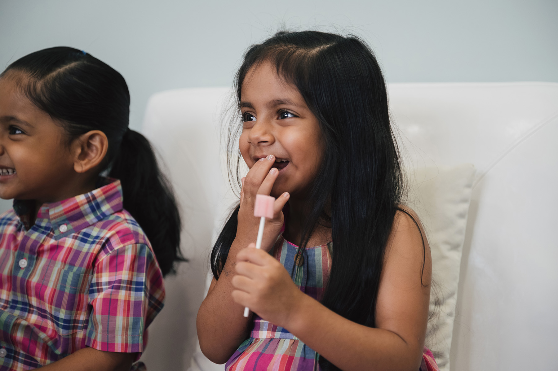Smiling girl with long black hair holding a pink lollipop while sitting on a white couch next to another child.