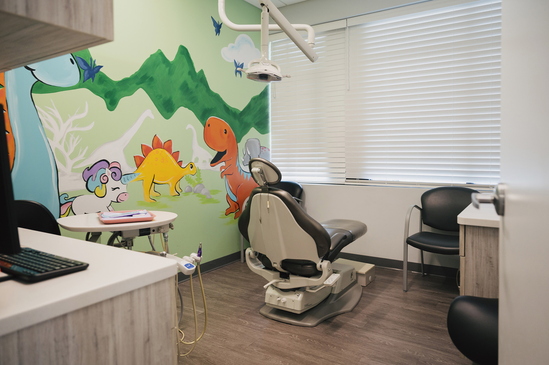 Children's dental exam room with a dental chair, equipment, and colorful dinosaur and unicorn mural on the wall.