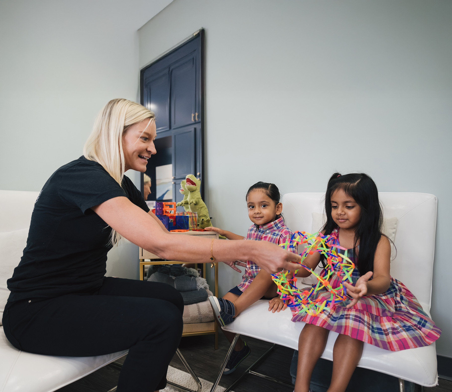 A woman interacts with two young children sitting on white chairs, one child holding a colorful expanding toy.