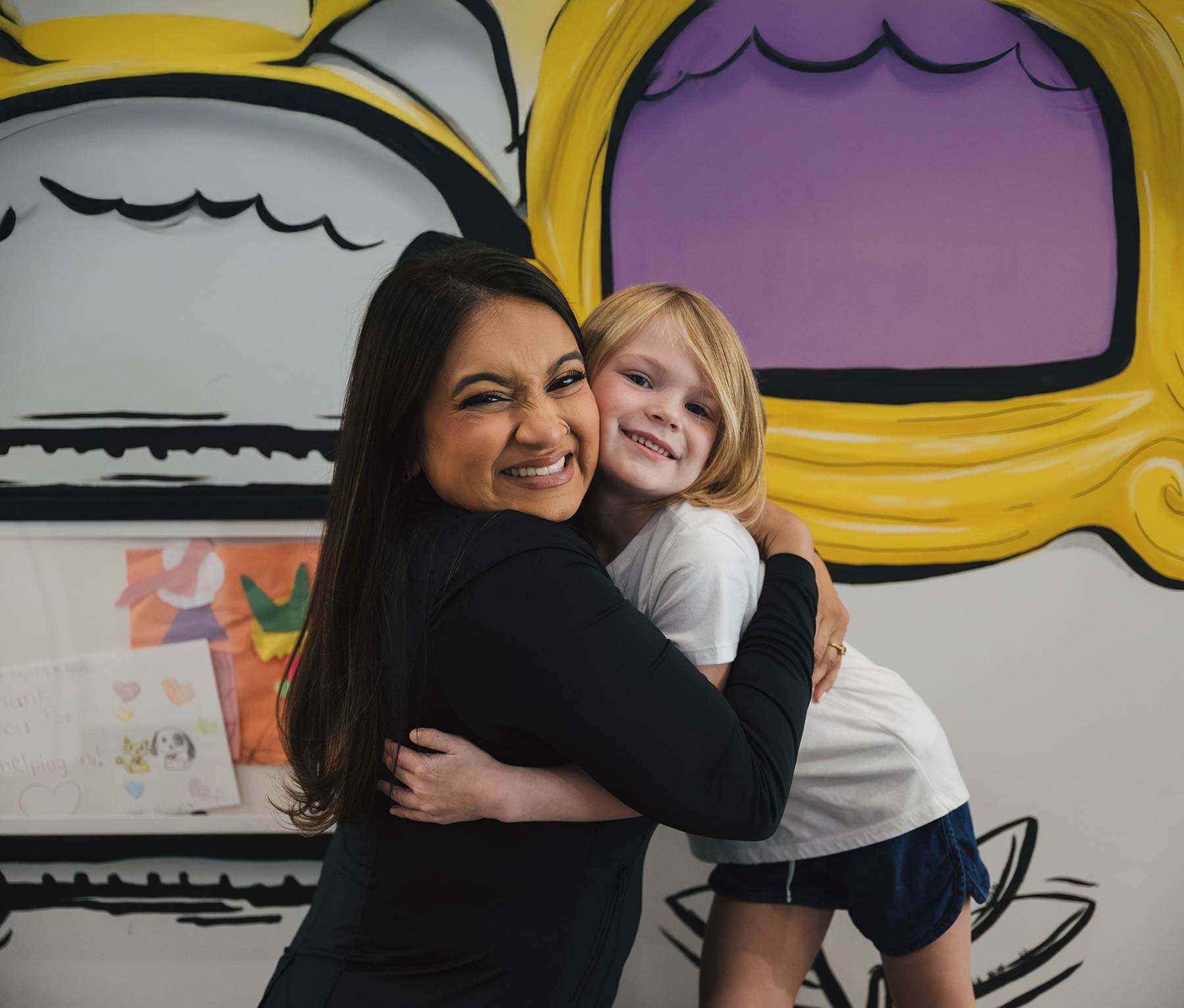 Smiling woman with long dark hair hugging a young blonde girl in front of a colorful mural.