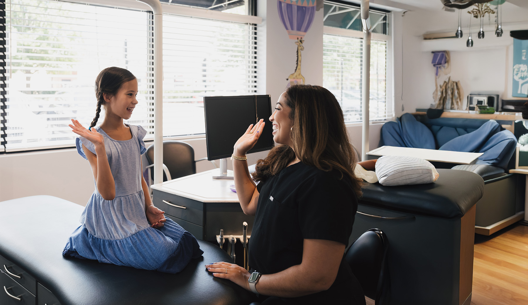 Young girl in a blue dress sitting on a medical examination table giving a high five to a smiling female healthcare professional in a clinic room.