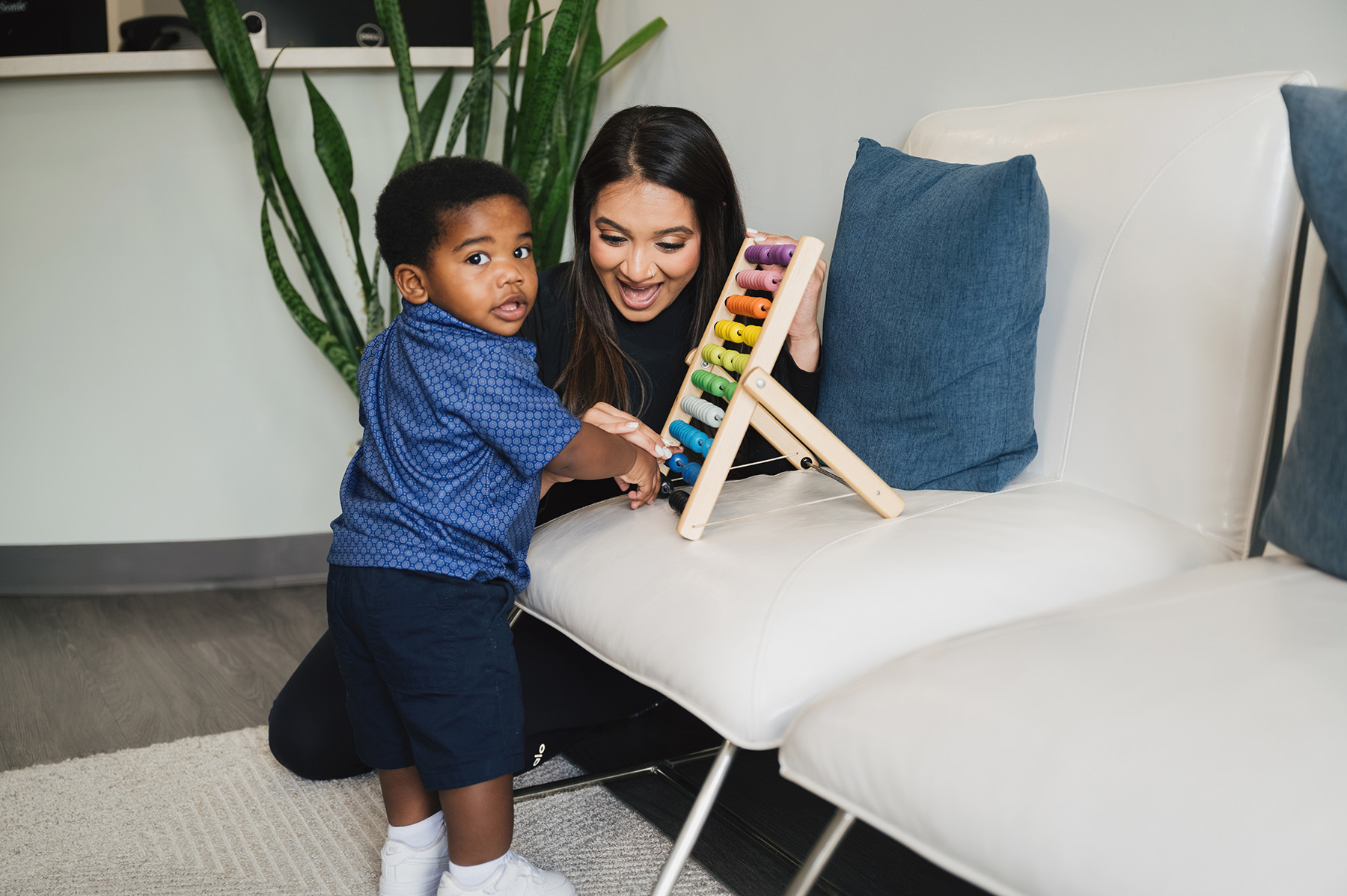 Woman kneeling and smiling while showing a colorful abacus to a toddler in a blue shirt and shorts standing by a white couch.