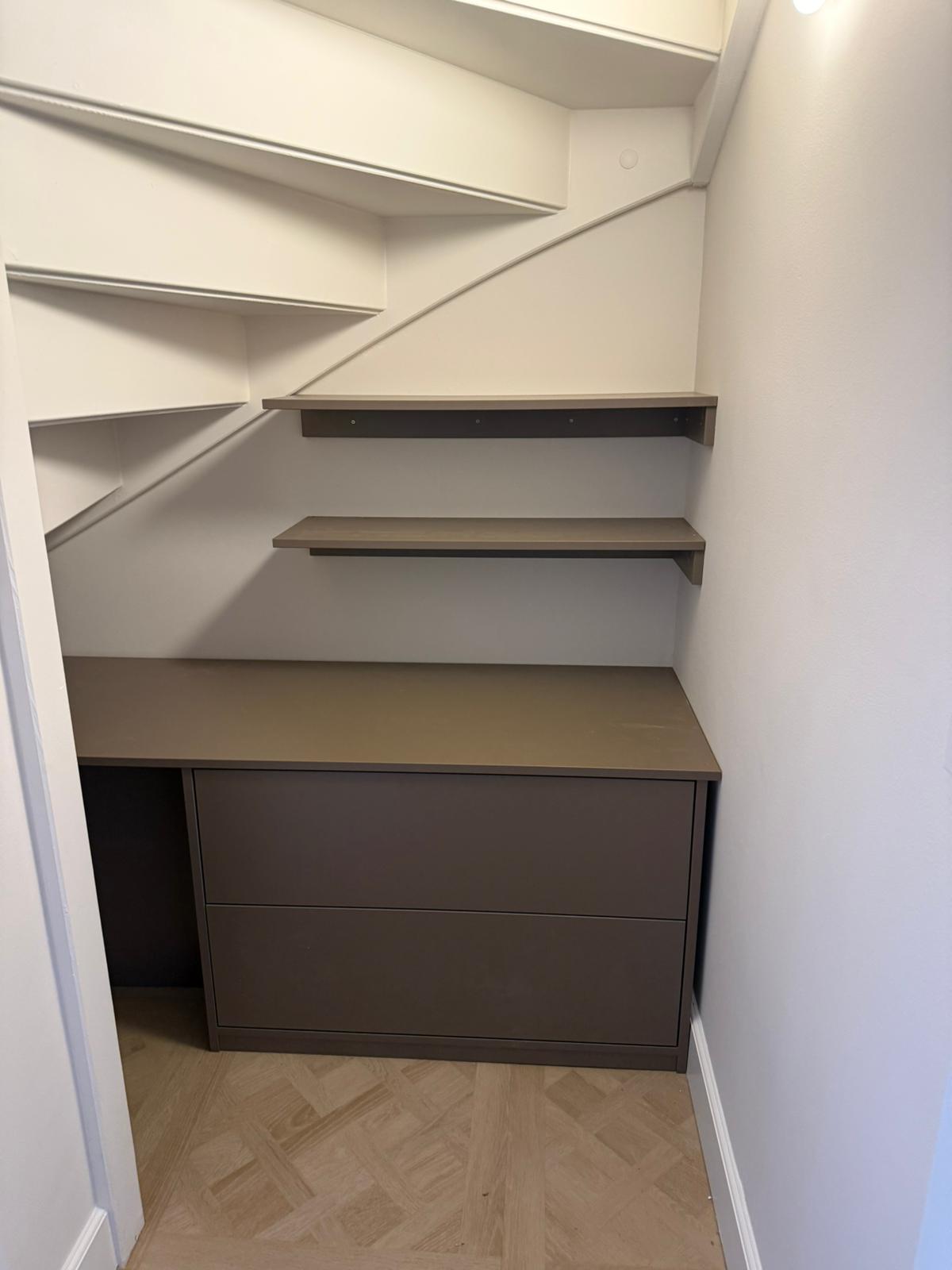 Lege opbergkast met twee lades en twee plankjes onder een witte trap in een kleine ruimte.