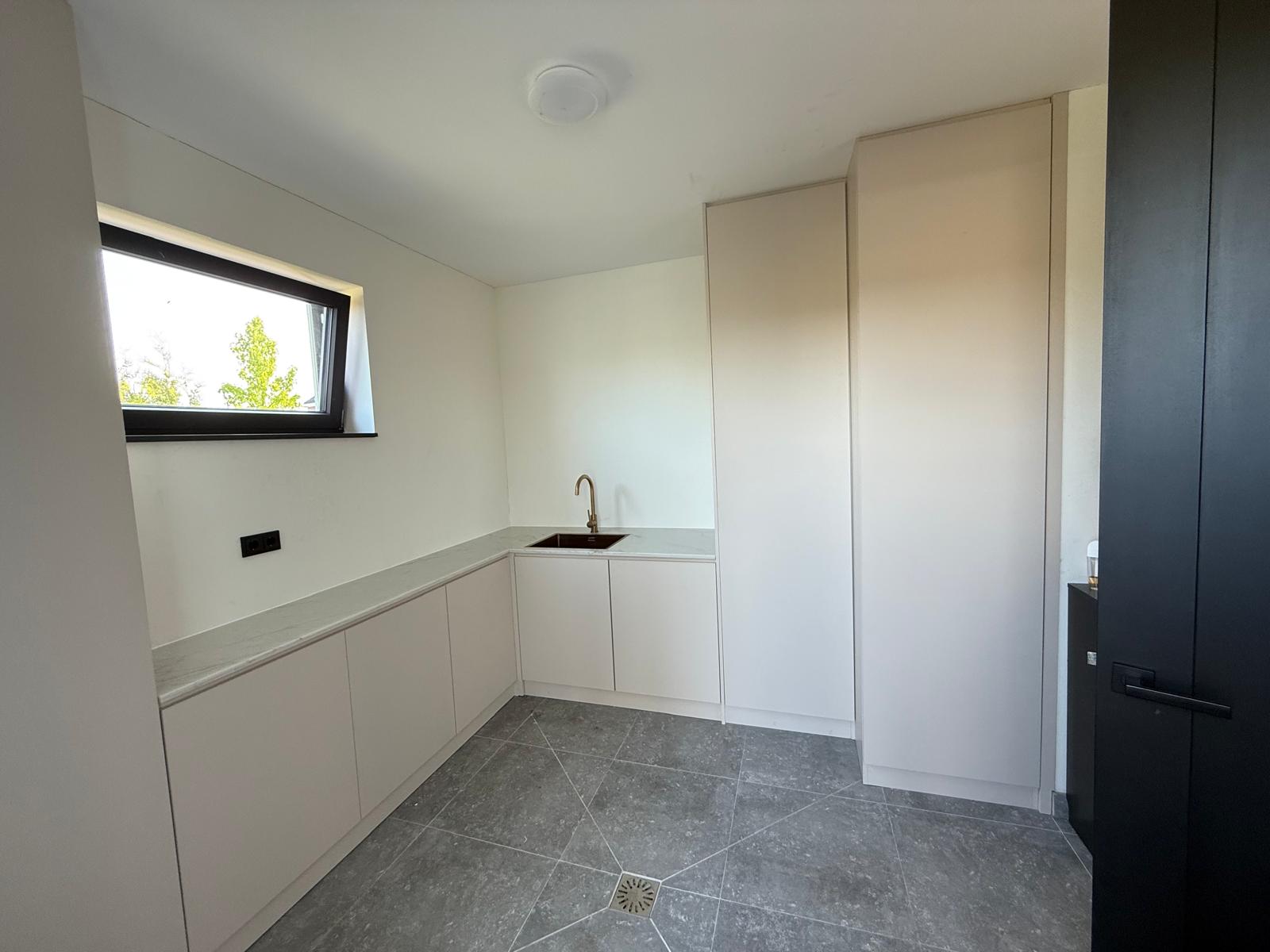 Moderne keuken met beige kastdeuren, grijs marmeren vloer, koperen kraan en een klein hoog raam met uitzicht op bomen.