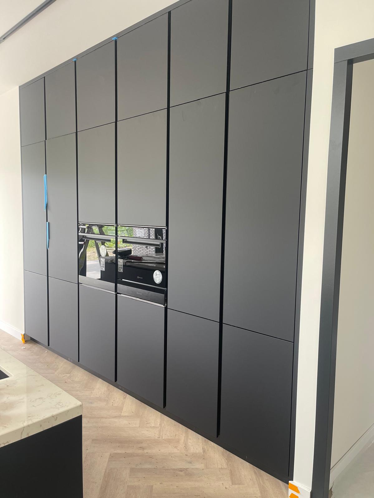 Moderne keukenwand met matzwarte kastdeuren en ingebouwde oven, licht houten visgraatvloer.