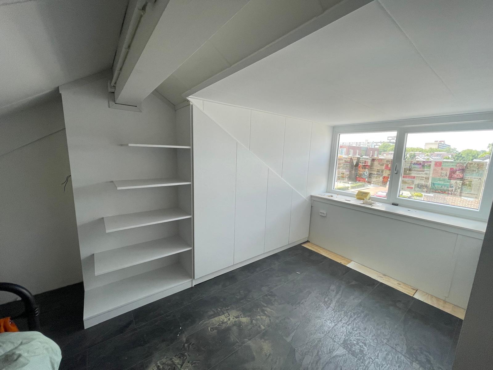 Lege kamer met zwarte tegelvloer, witte ingebouwde kast en plankjes naast een raam met zicht op buiten.