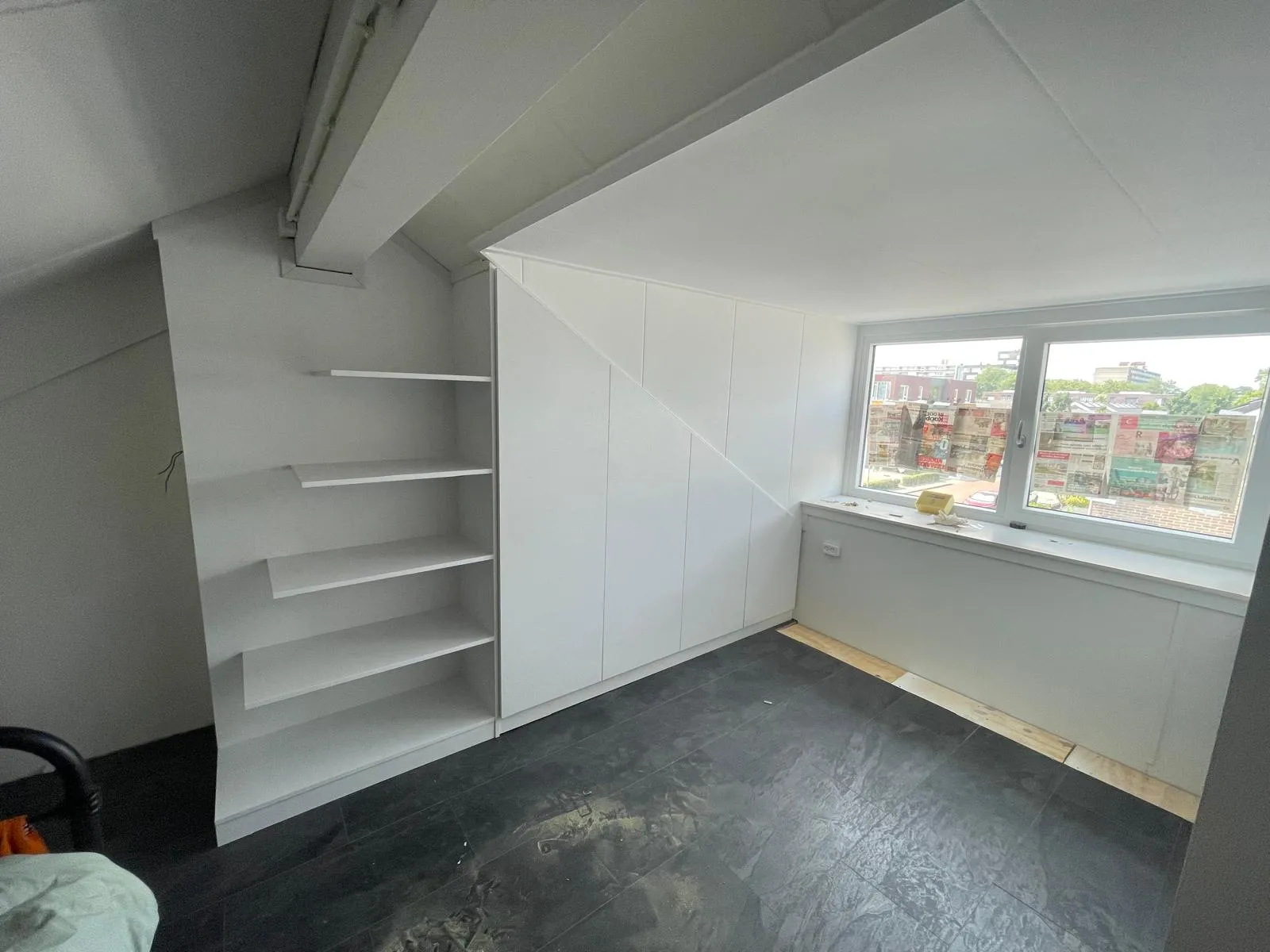 Lege kamer met zwarte tegelvloer, witte ingebouwde kast en plankjes naast een raam met zicht op buiten.