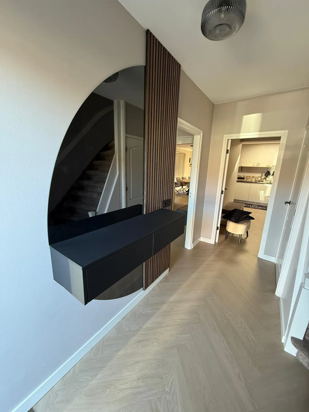 Moderne hal met lichte houten vloer, spiegel met zwart zwevend kastje aan de muur en doorgang naar een woonkamer en keuken.