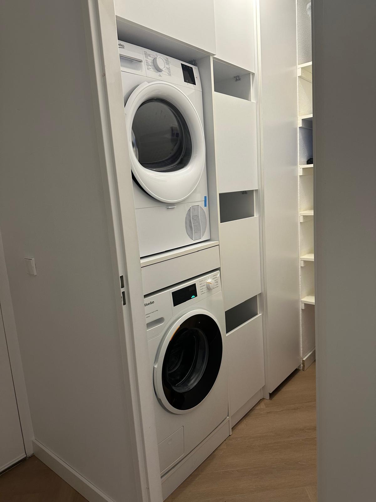 Wasmachine en droger gestapeld in een smalle wasruimte met witte kastdeuren en houten vloer.