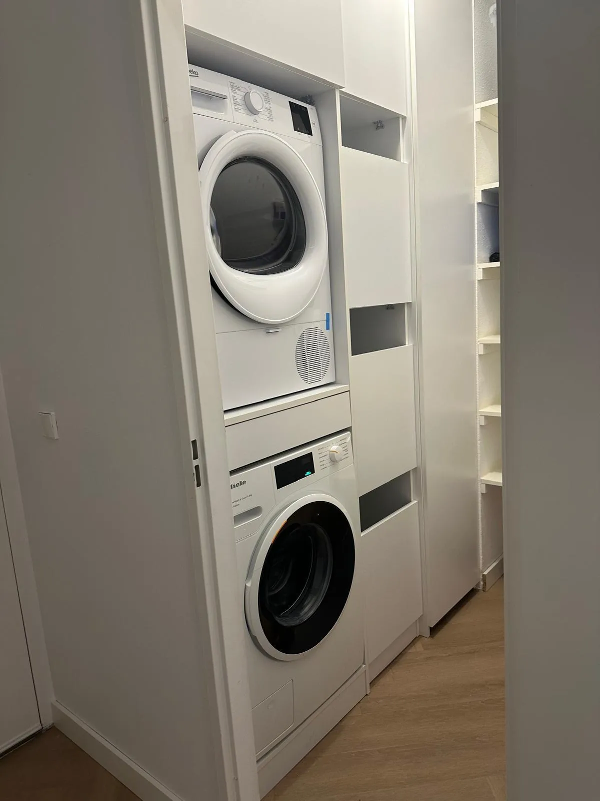Wasmachine en droger gestapeld in een smalle wasruimte met witte kastdeuren en houten vloer.