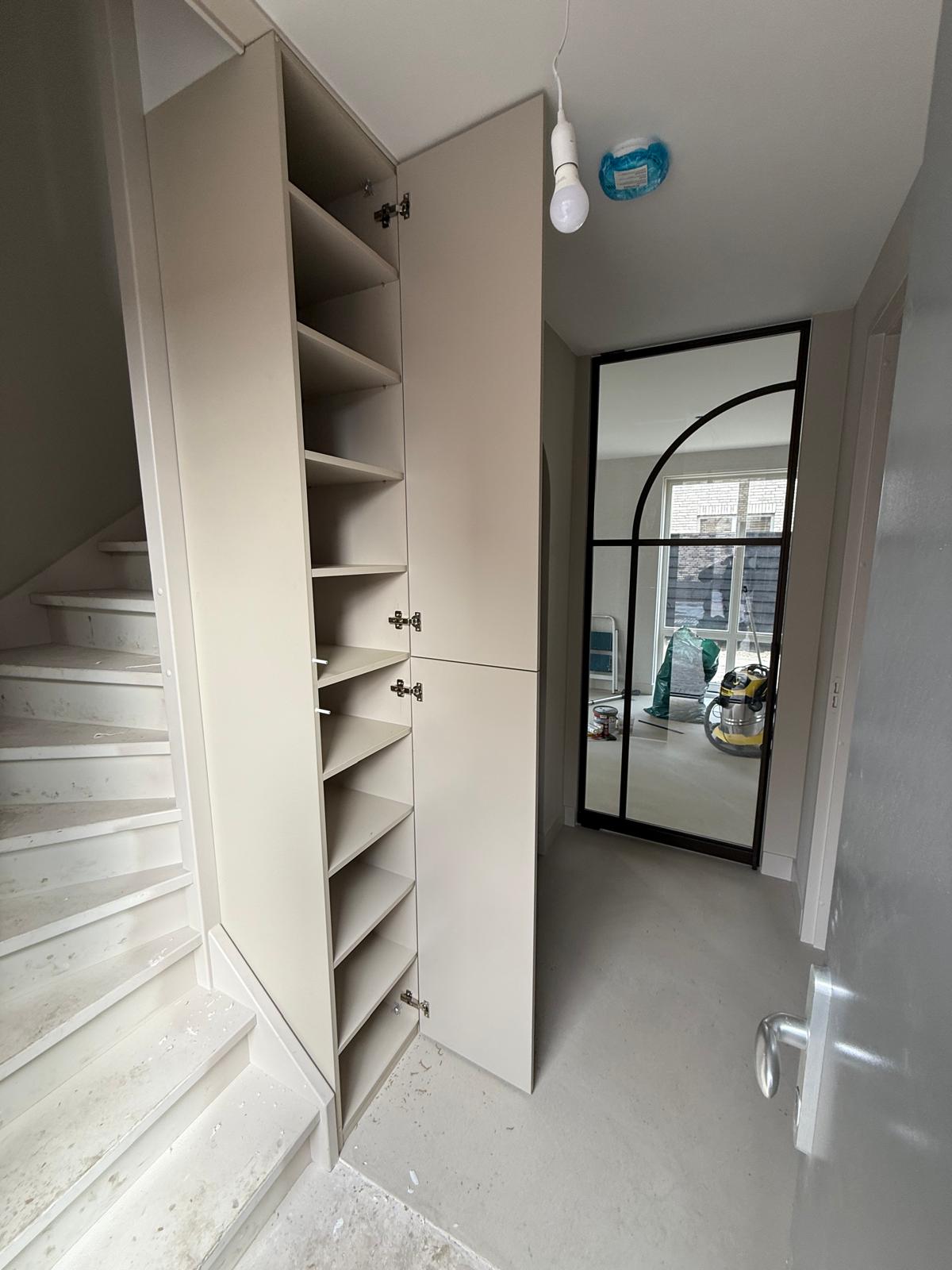 Lege beige kast met open deuren naast een witte trap in een hal met een glazen deur met zwart metalen frame en bouwmaterialen op de achtergrond.