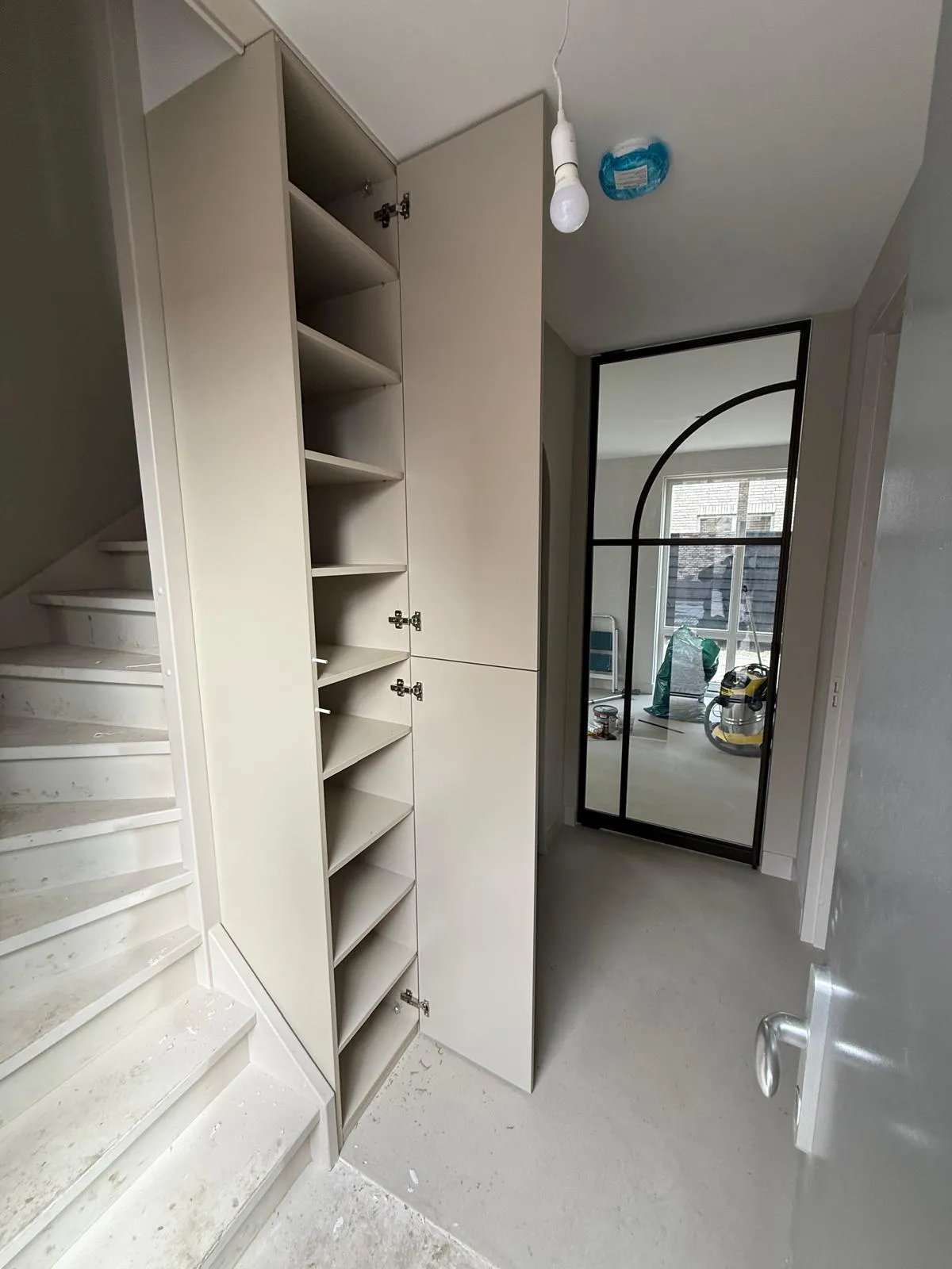 Lege beige kast met open deuren naast een witte trap in een hal met een glazen deur met zwart metalen frame en bouwmaterialen op de achtergrond.