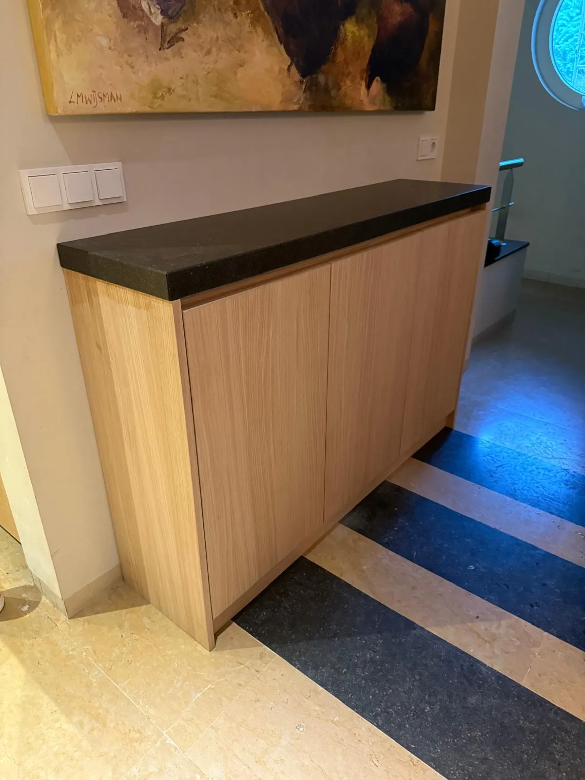 Moderne houten kast met een zwart granieten blad in een gang met een geschilderd kunstwerk aan de muur.