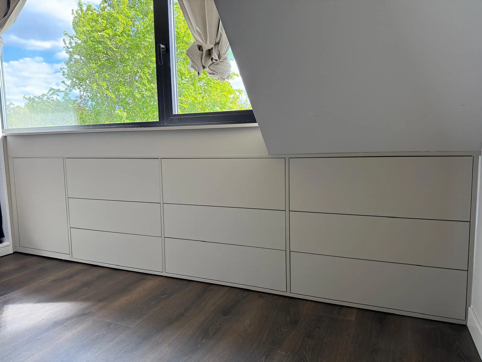 Lage ingebouwde kasten onder een schuin plafond met een raam erboven en donkere houten vloer.