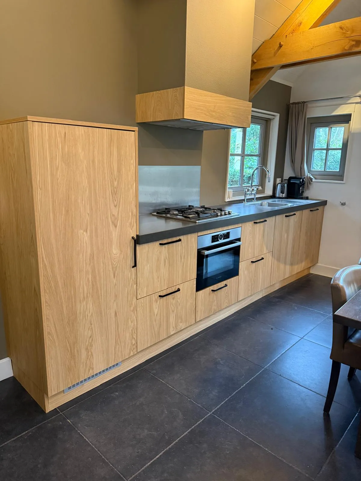 Moderne keuken met houten kasten, een ingebouwde oven, gaskookplaat, dubbele gootsteen en groot zwart werkblad onder twee ramen.