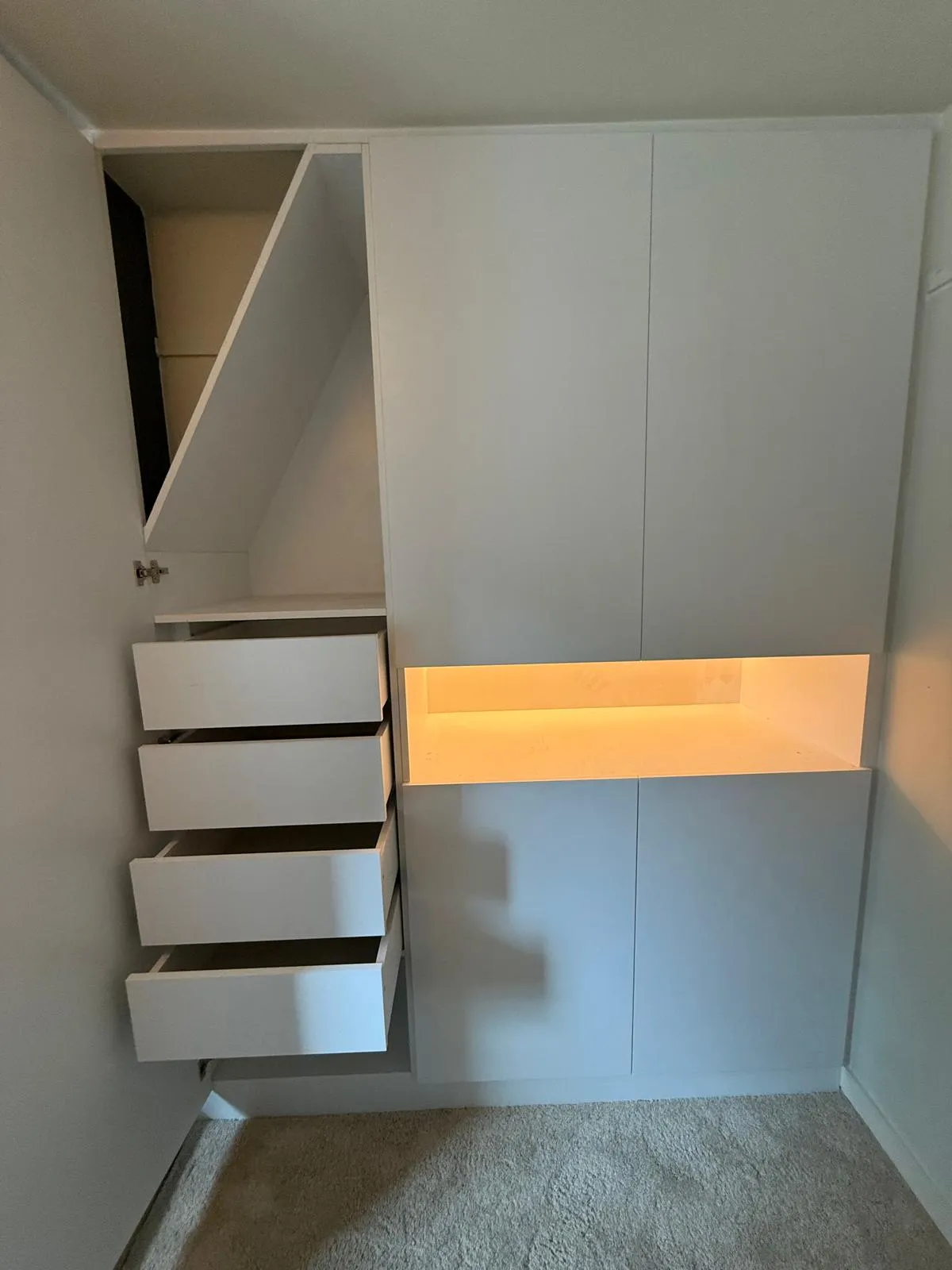 Witte kastenwand met vier open lades links, dichte kastdeuren rechts, en een verlicht nisje in het midden.