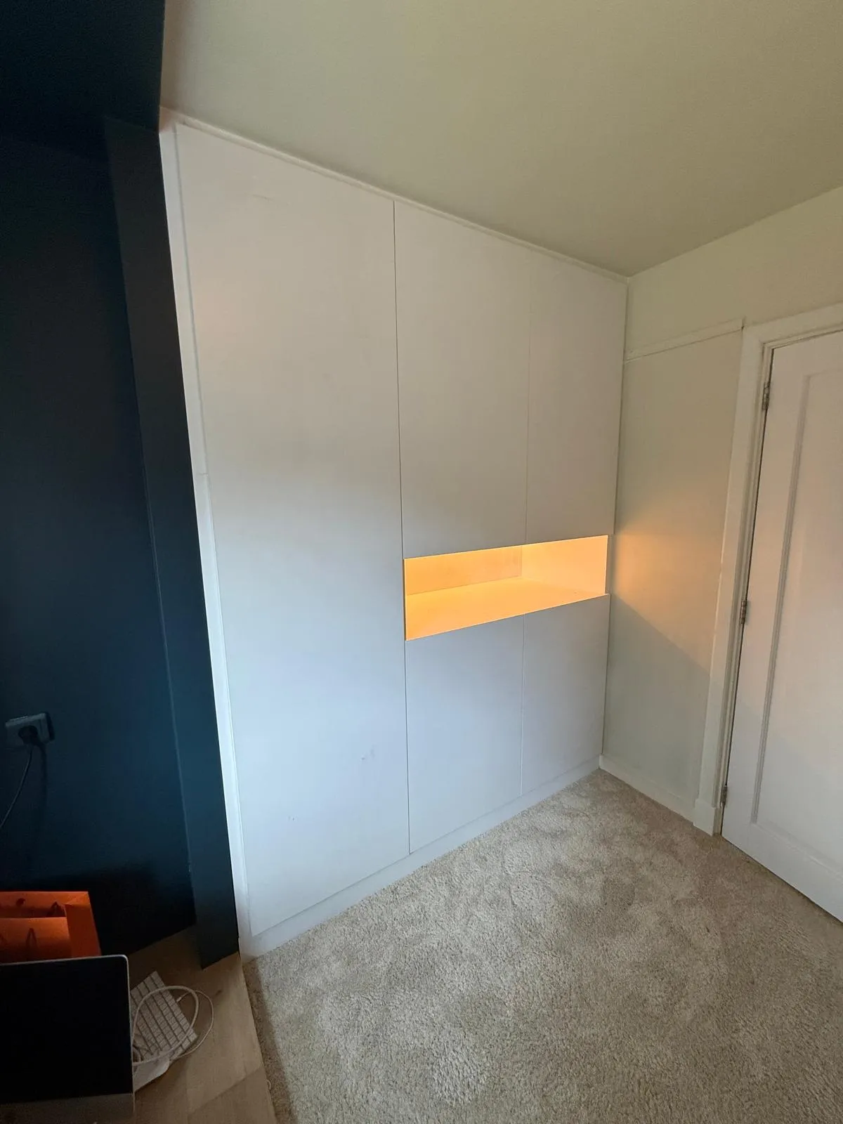 Witte ingebouwde kast met centrale verlichte open plank in een kamer met grijs tapijt en een wit deur.