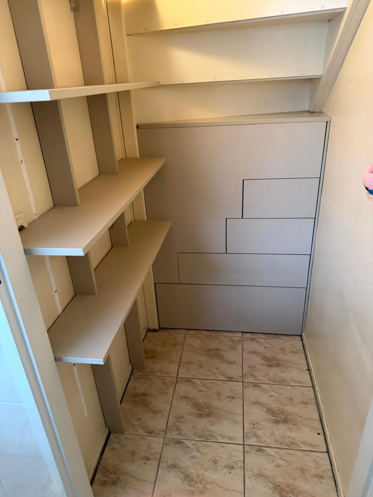 Kleine opslagruimte met drie lage witte planken aan de linkerwand en een kast met trapdeur aan de achterkant.