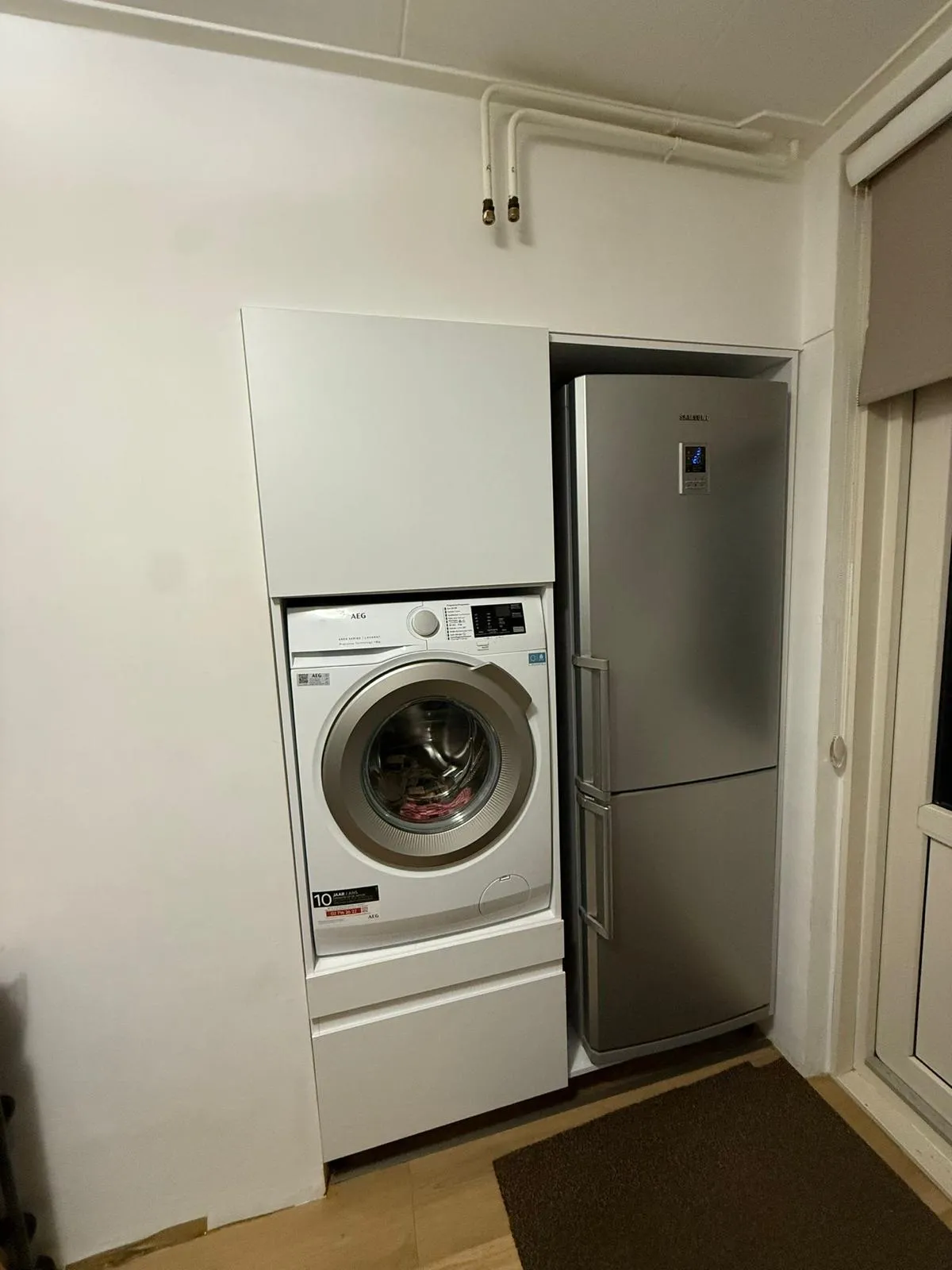 Ingebouwde witte AEG wasmachine naast een roestvrijstalen koelkast in een keukenruimte met houten vloer en een bruine mat.