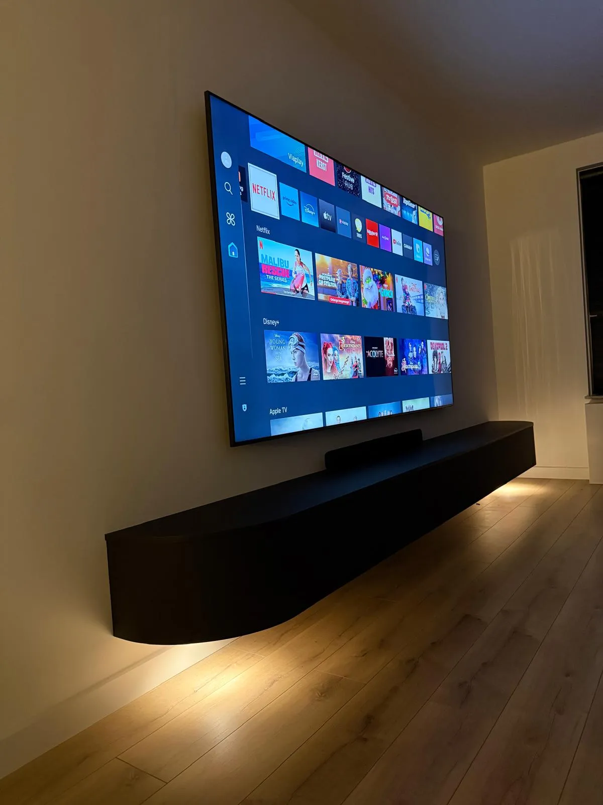 Wandgemonteerde televisie met streamingapps zoals Netflix, Disney+ en Apple TV op het scherm boven een zwevende zwarte plank met sfeerverlichting.