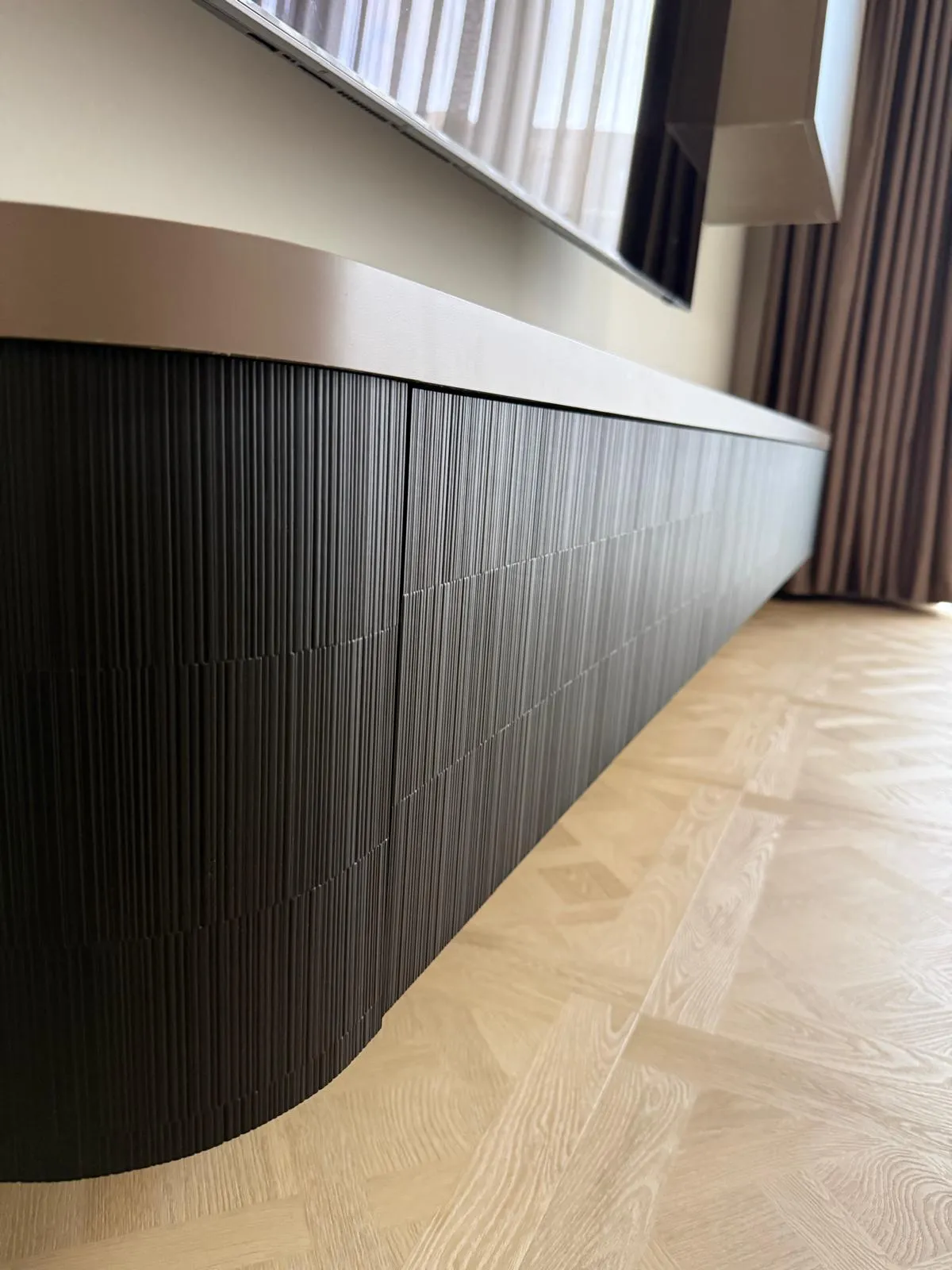 Modern woonkamer met een grote vlakke muurbeugeltelevisie boven een laag zwart kastmeubel met verticale lijnen en een houten vloer met patroon.