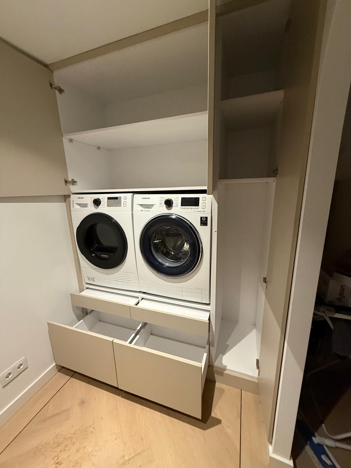 Wasmachine en droger geïntegreerd in een kast met open deuren en laden, in een wasruimte.