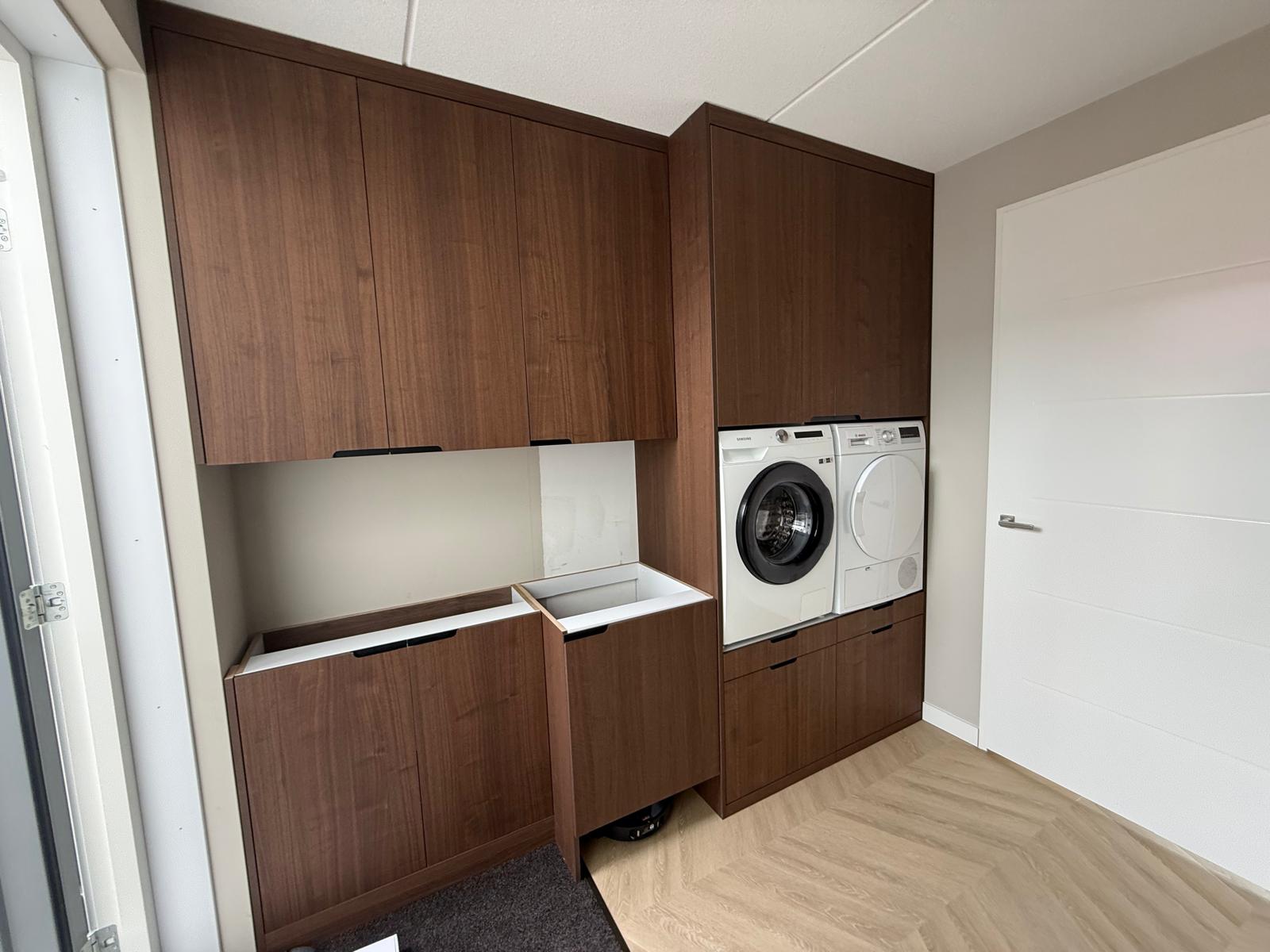 Moderne wasruimte met ingebouwde wasmachine, droger en houten kastdeuren, lichte vloer en witte deur.