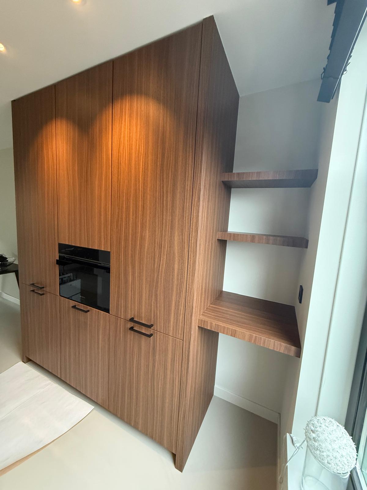 Moderne houten kast met ingebouwde oven en open planken aan de rechterkant bij een raam.
