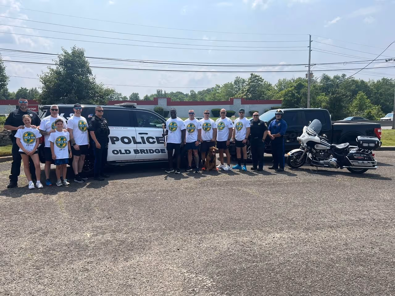 A group of people standing in front of a parked police vehicle under an open sky, surrounded by trees and on a road.