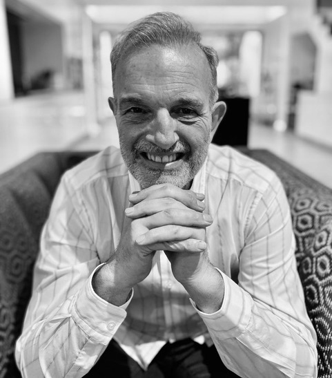Smiling middle-aged man with gray hair and beard sitting on a patterned couch, hands clasped near his face.