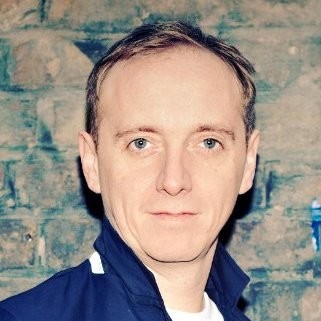 Portrait of a man with short light brown hair wearing a blue jacket against a brick wall background.