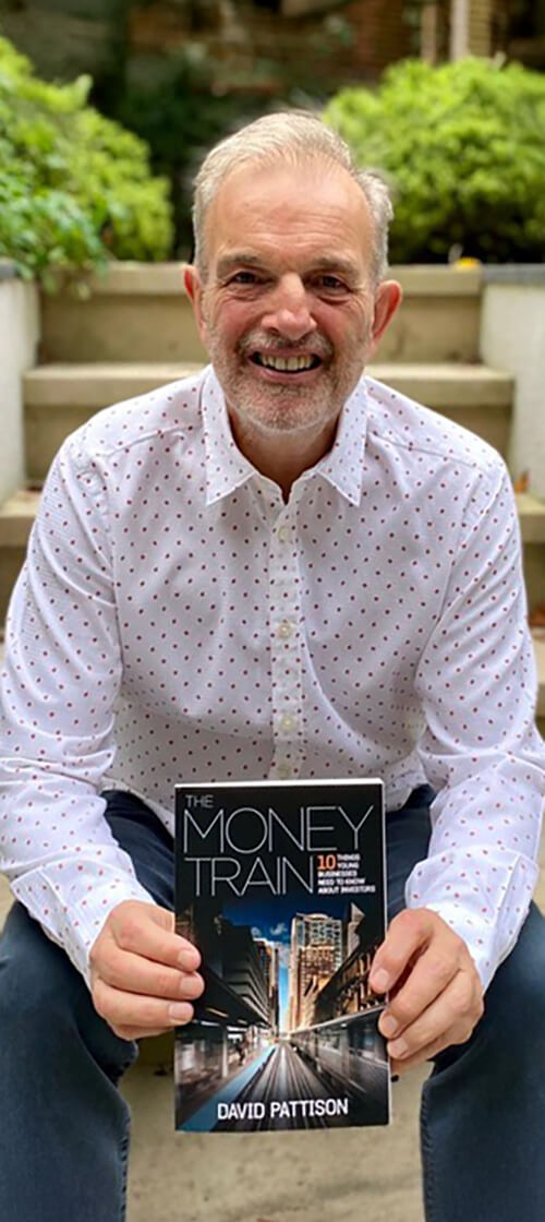 Smiling man in a white shirt with small red dots sitting outdoors and holding a book titled 'The Money Train' by David Pattison.